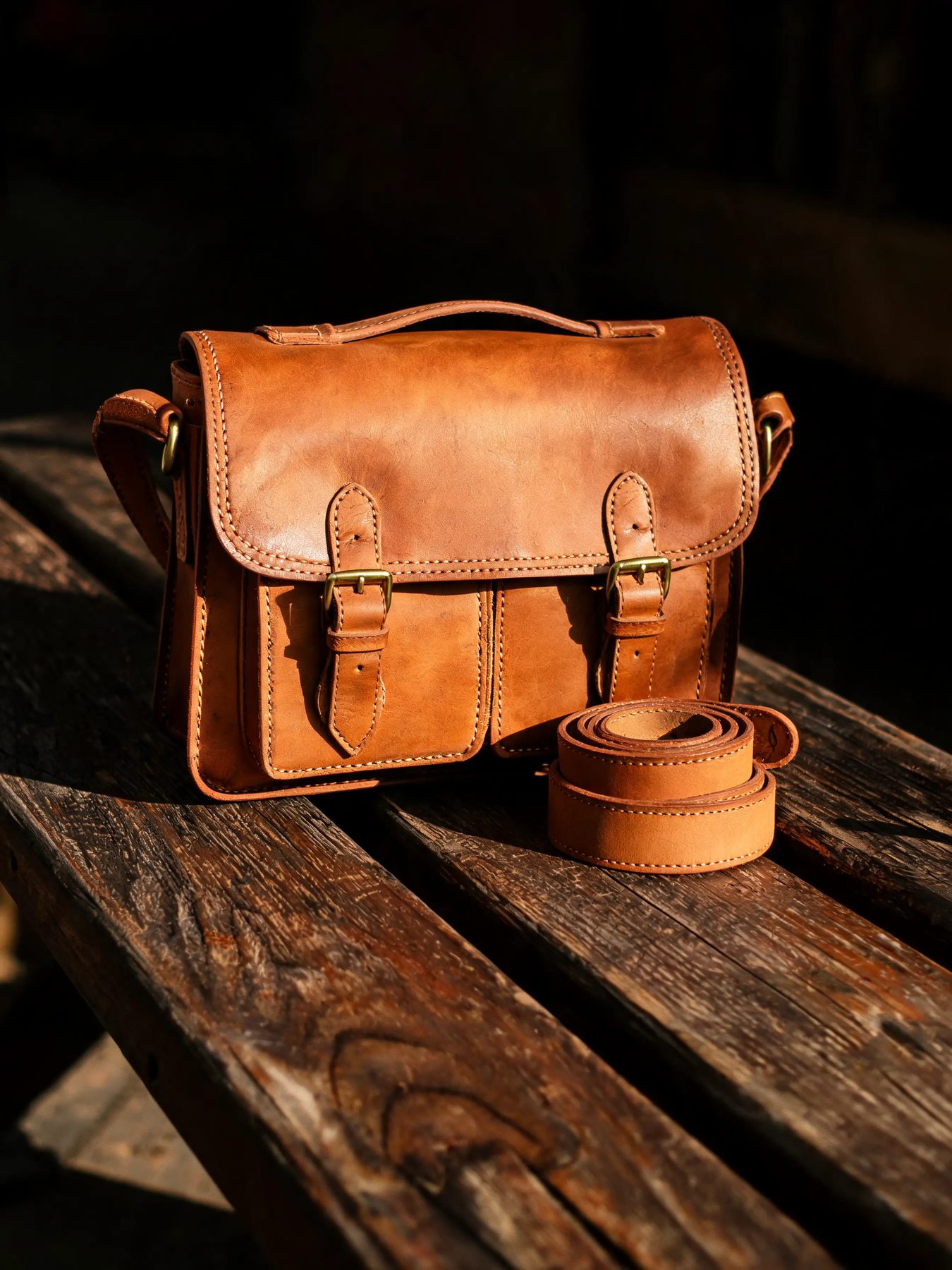 Vegetable-tanned leather satchel with brass buckles beside a coiled belt on a wooden bench